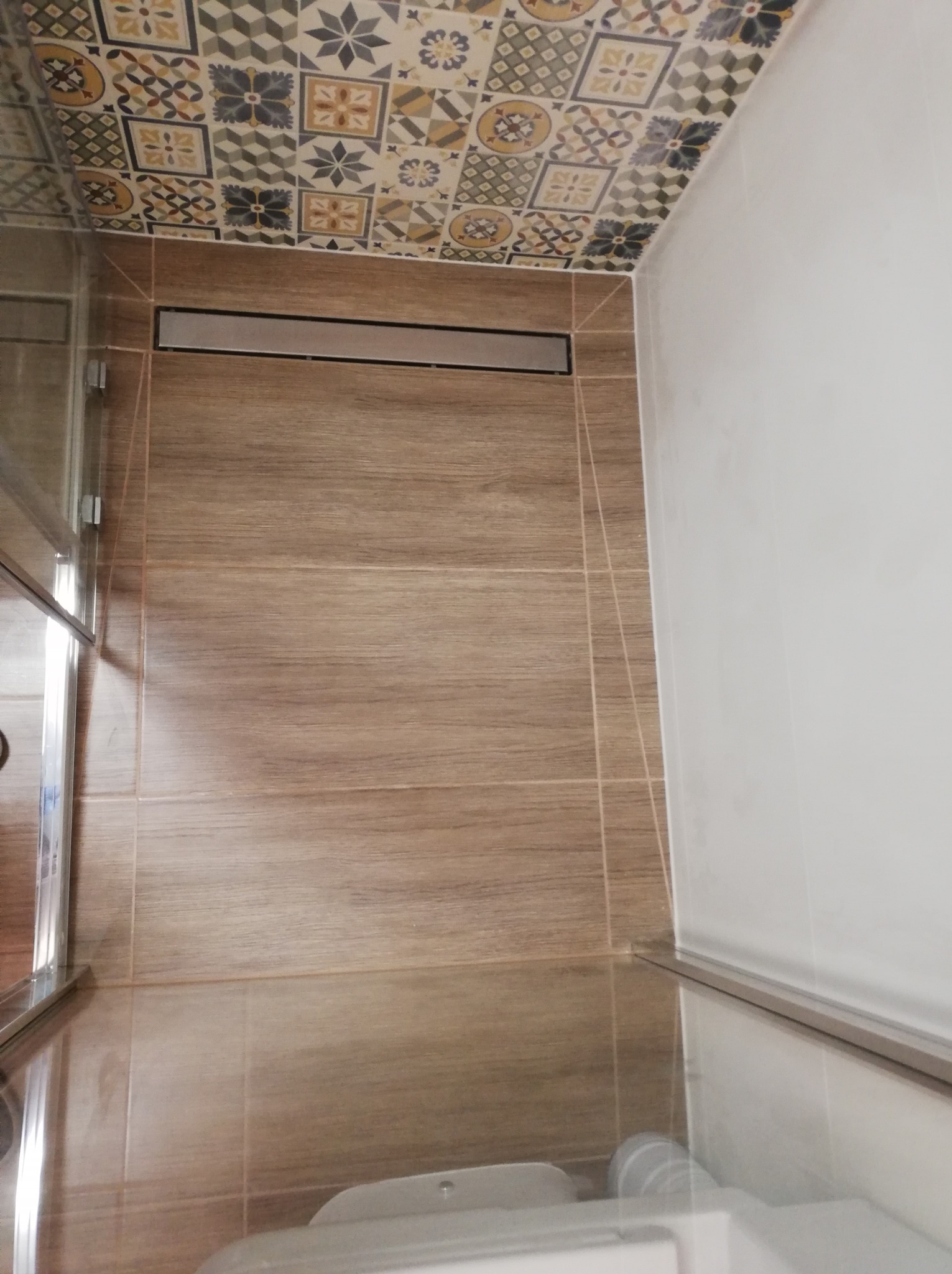 Wykończona kabina prysznicowa z odpływem liniowym, brązowe płytki imitujące drewno na ścianach i podłodze, sufit wykończony dekoracyjnymi płytkami w stylu patchwork.