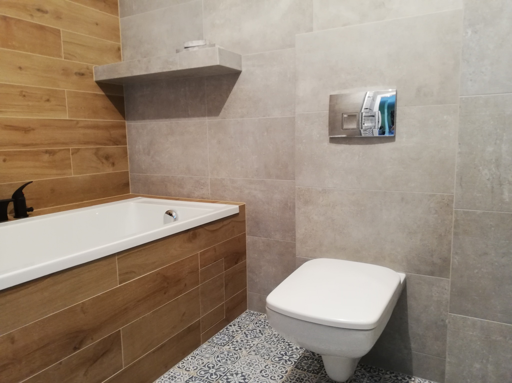 Nowoczesna łazienka po remoncie: brązowe płytki imitujące drewno przy wannie, szare płytki na ścianach, podwieszana toaleta i dekoracyjne płytki podłogowe.