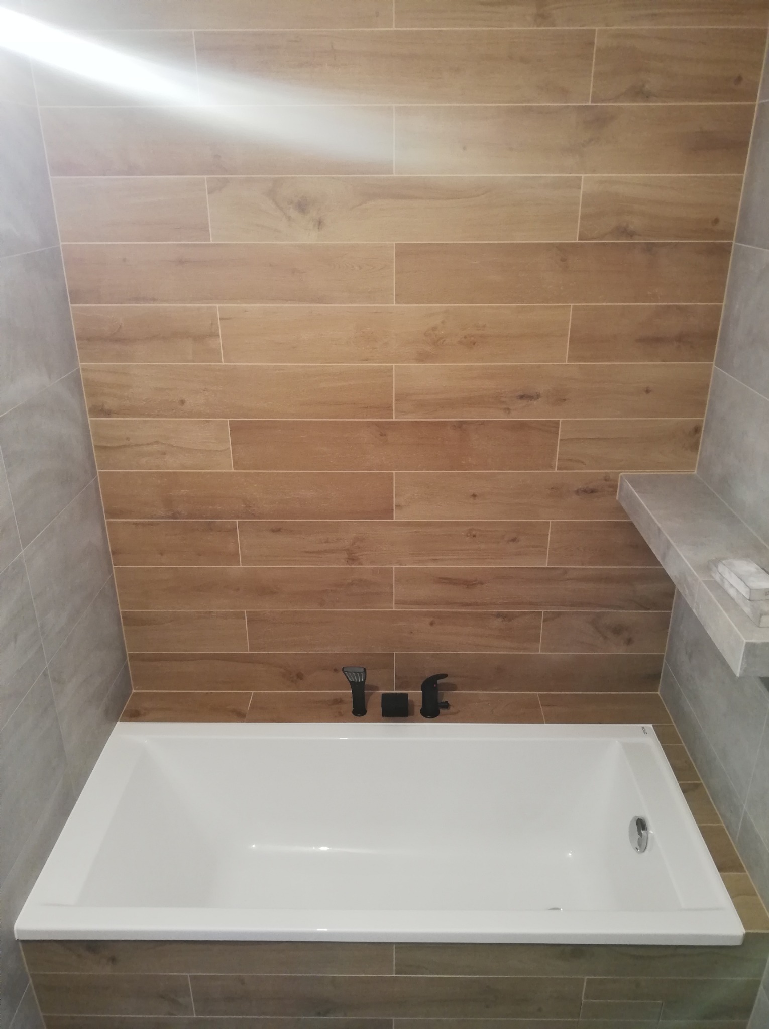 Nowoczesna łazienka po remoncie: biała wanna obudowana płytkami imitującymi drewno, ściany wyłożone płytkami w odcieniach szarości, czarna bateria wannowa.