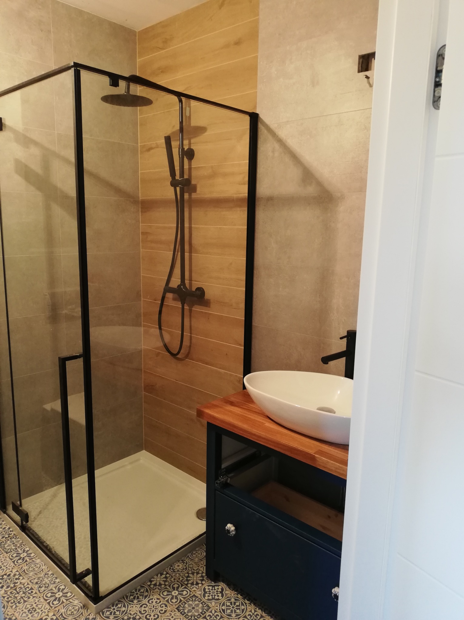 Nowoczesna łazienka z kabiną prysznicową typu walk-in z czarnymi profilami, umywalką nablatową na drewnianym blacie i szafką w kolorze granatowym oraz wzorzystą podłogą.