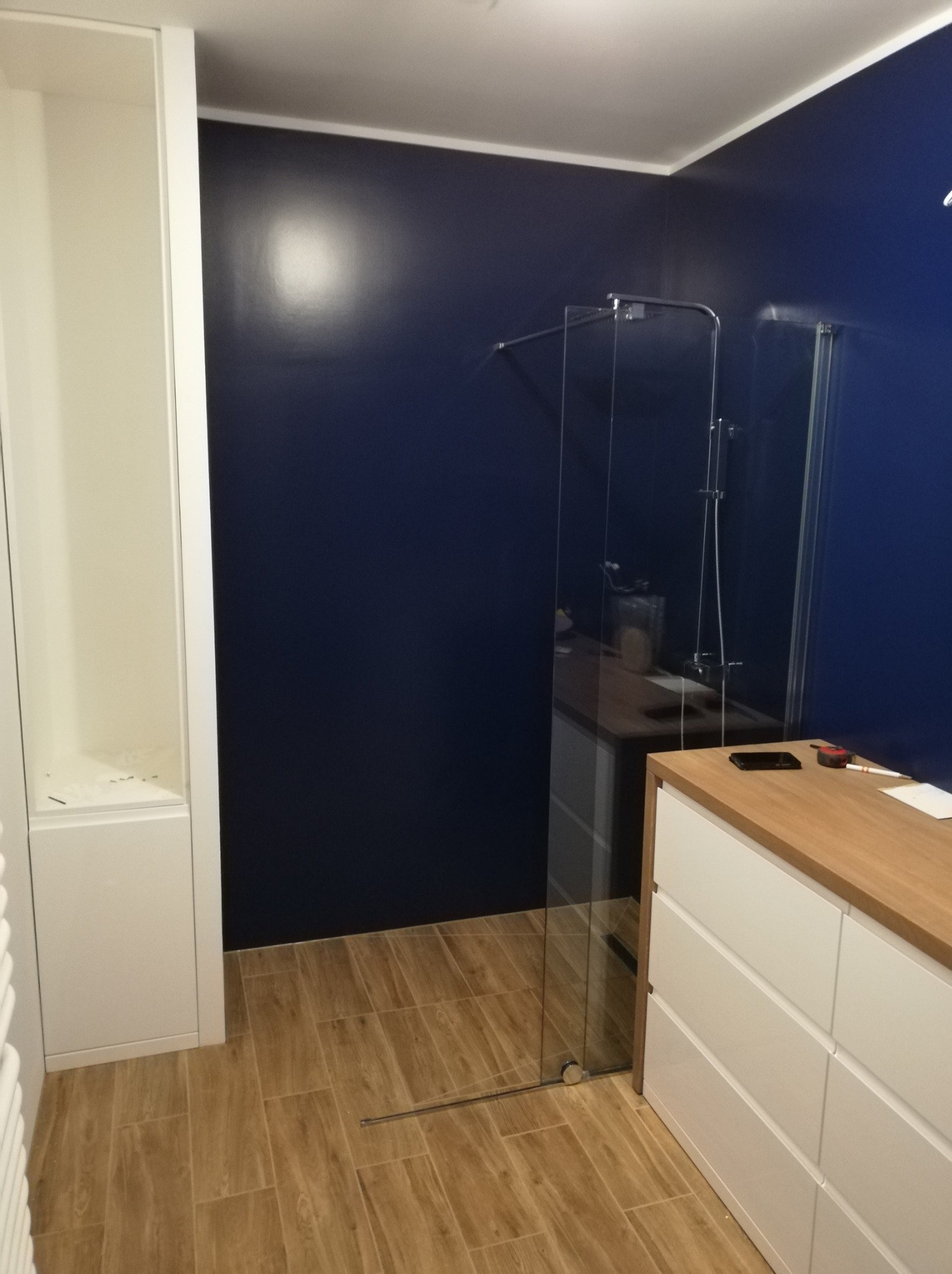 Nowoczesna łazienka z ciemnoniebieskimi ścianami, kabiną prysznicową ze szkła, drewnopodobną podłogą, białą szafką z szufladami i otwartą szafką w zabudowie.