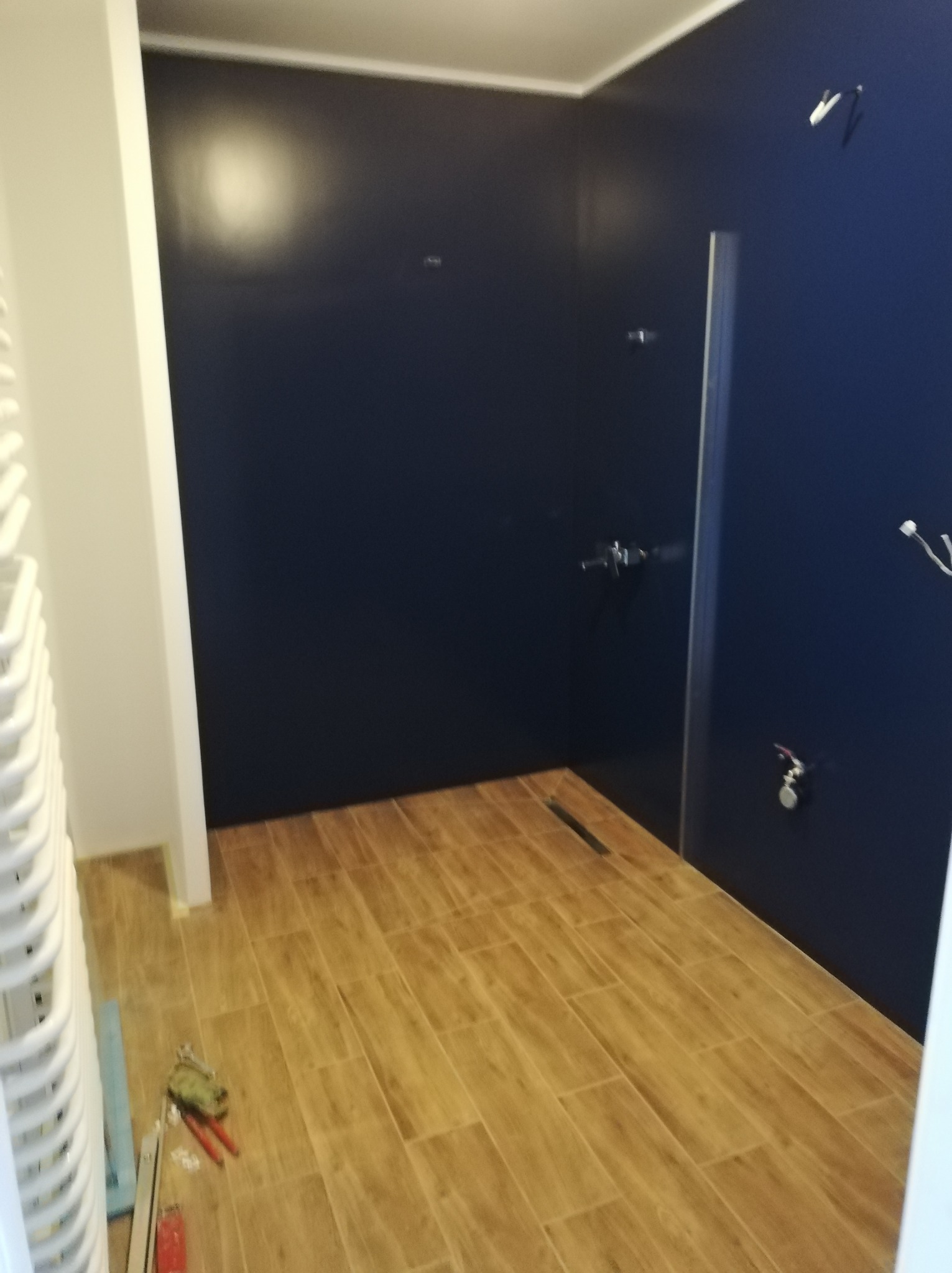 Niedokończona łazienka z ciemnymi ścianami, drewnopodobną podłogą, grzejnikiem drabinkowym i narzędziami na podłodze.