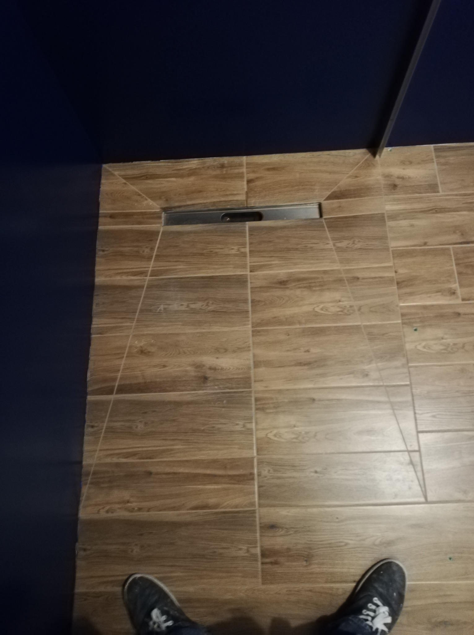 Ułożone płytki podłogowe imitujące drewno w łazience, widoczny odpływ liniowy i fragment niebieskiej ściany oraz stopy w butach.