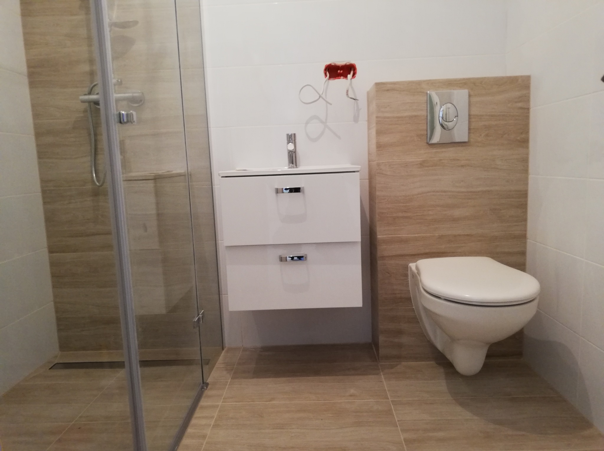 Nowoczesna łazienka z kabiną prysznicową, wiszącą szafką z umywalką i podwieszanym WC, wykończona płytkami imitującymi drewno, z widoczną puszką elektryczną w ścianie.