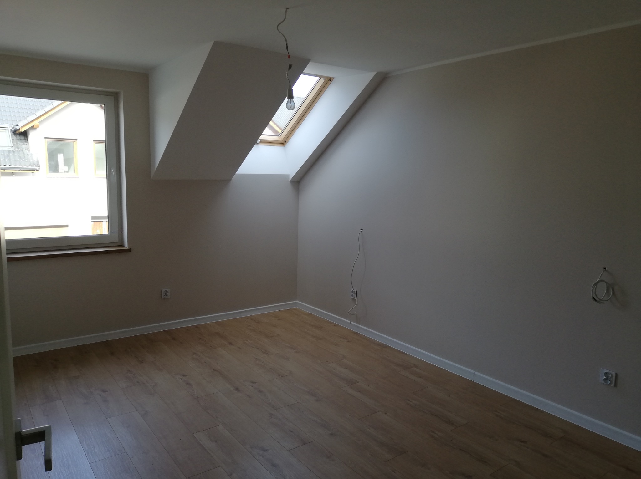 Puste, jasne poddasze z oknem dachowym, panele podłogowe w odcieniach brązu, białe ściany przygotowane do malowania, widoczne gniazdka elektryczne i wystające kable.