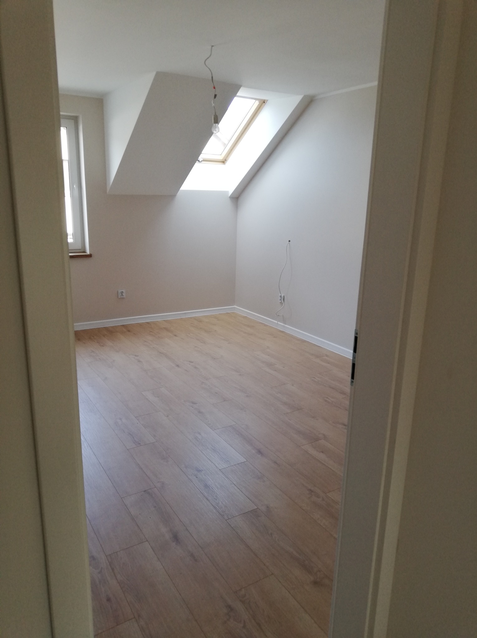 Poddasze z oknem dachowym po remoncie, świeżo ułożone panele podłogowe w odcieniach brązu, białe ściany i listwy przypodłogowe, widok zza futryny drzwi.