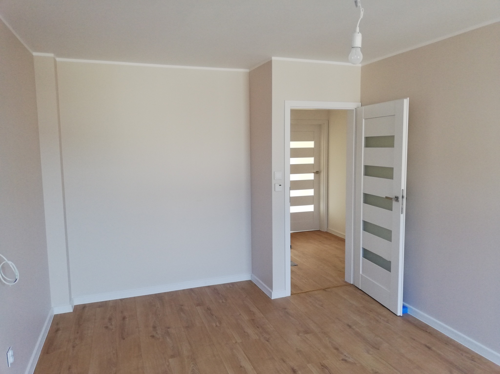 Pomalowane na beżowo wnętrze pokoju z panelami podłogowymi, białymi listwami przypodłogowymi, otwartymi białymi drzwiami z przeszkleniami i wiszącą żarówką.