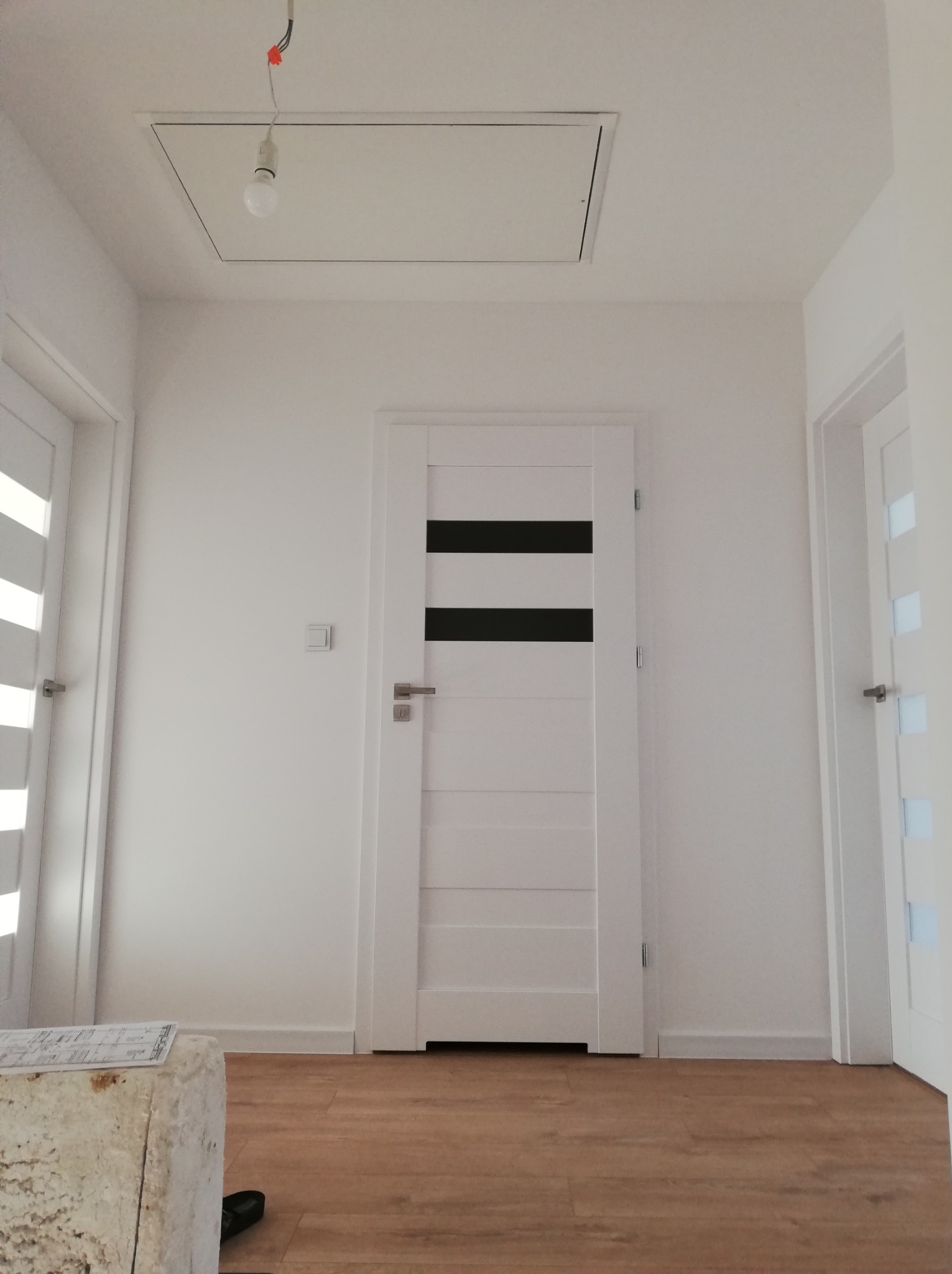 Nowoczesny korytarz po remoncie: białe ściany, panele podłogowe, dwie białe drzwi z czarnymi i przezroczystymi wstawkami, sufit z klapą rewizyjną i wiszącą żarówką.