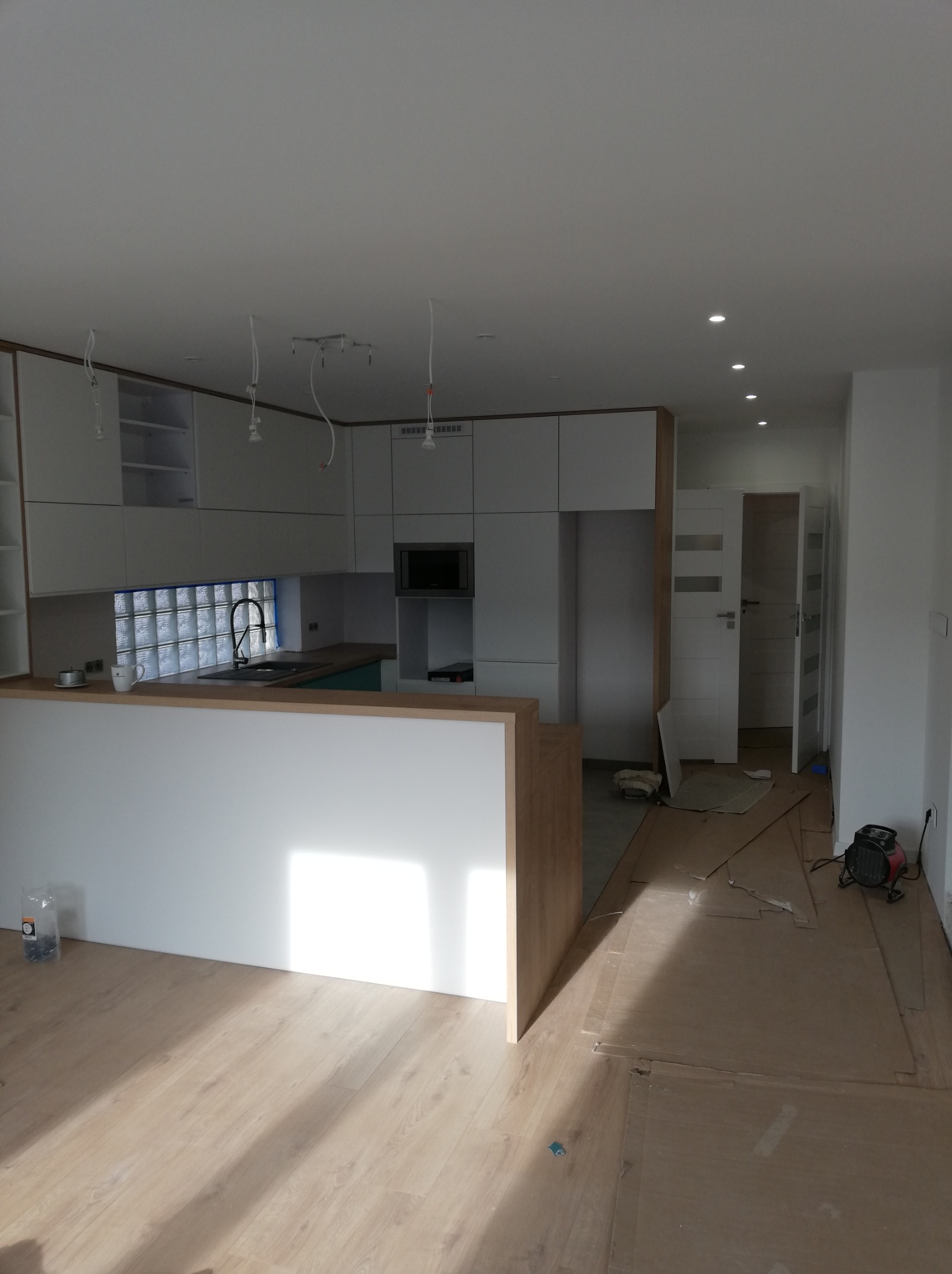 Wnętrze mieszkania w trakcie prac wykończeniowych, widoczna kuchnia z wyspą, częściowo ułożona podłoga, wiszące kable od oświetlenia i grzejnik elektryczny.