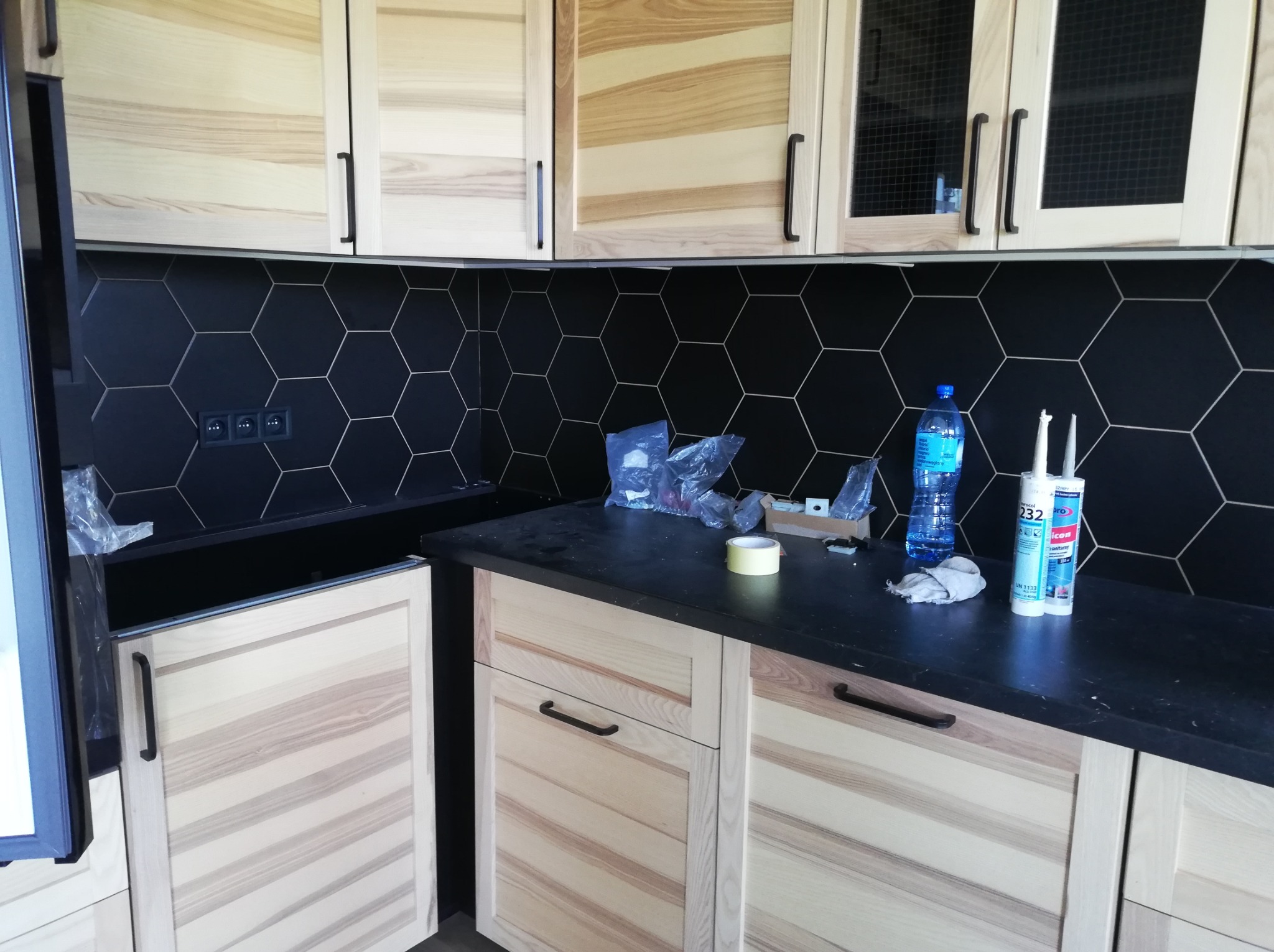 Częściowo ukończona zabudowa kuchenna z drewnianymi frontami, czarnym blatem i heksagonalnymi płytkami na ścianie, widoczne silikony i butelka wody na blacie.