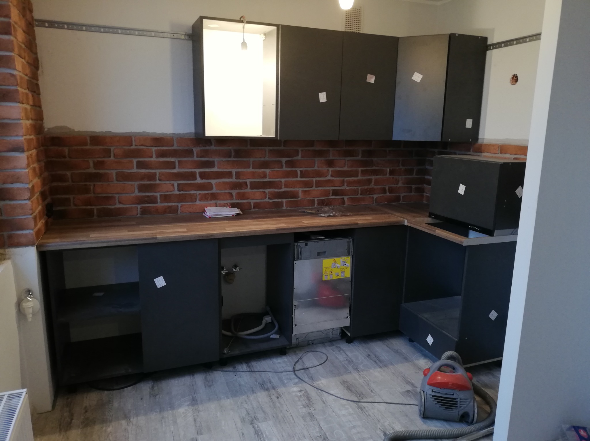 Montaż szarych szafek kuchennych z drewnianym blatem i ceglaną ścianą, widoczny podłączony sprzęt AGD i odkurzacz na podłodze.