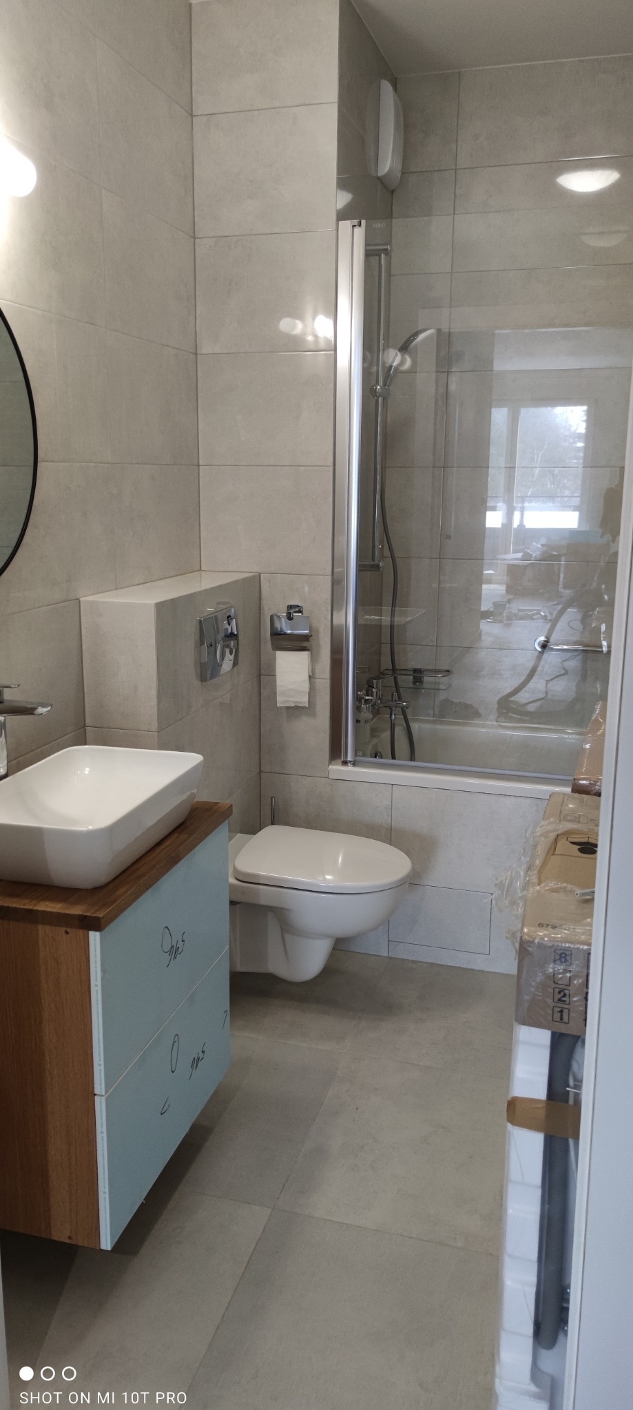 Ukończona łazienka z szarymi płytkami, wiszącą toaletą, umywalką nablatową na szafce z drewnianym blatem i kabiną prysznicową ze szklanymi drzwiami. W tle widoczne okno.