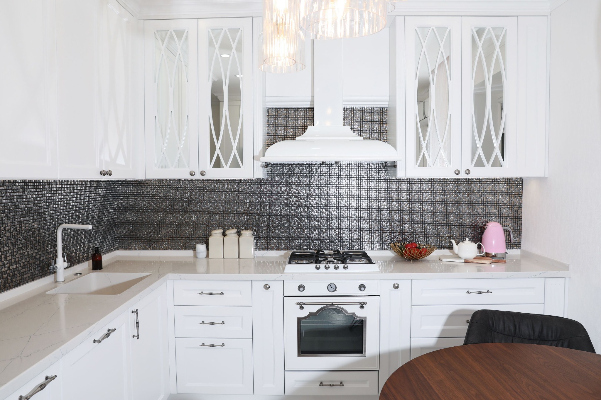 Biała kuchnia z błyszczącą mozaiką na ścianie, szafki z przeszklonymi frontami, okrągły drewniany stół i czarne krzesło w rogu.
