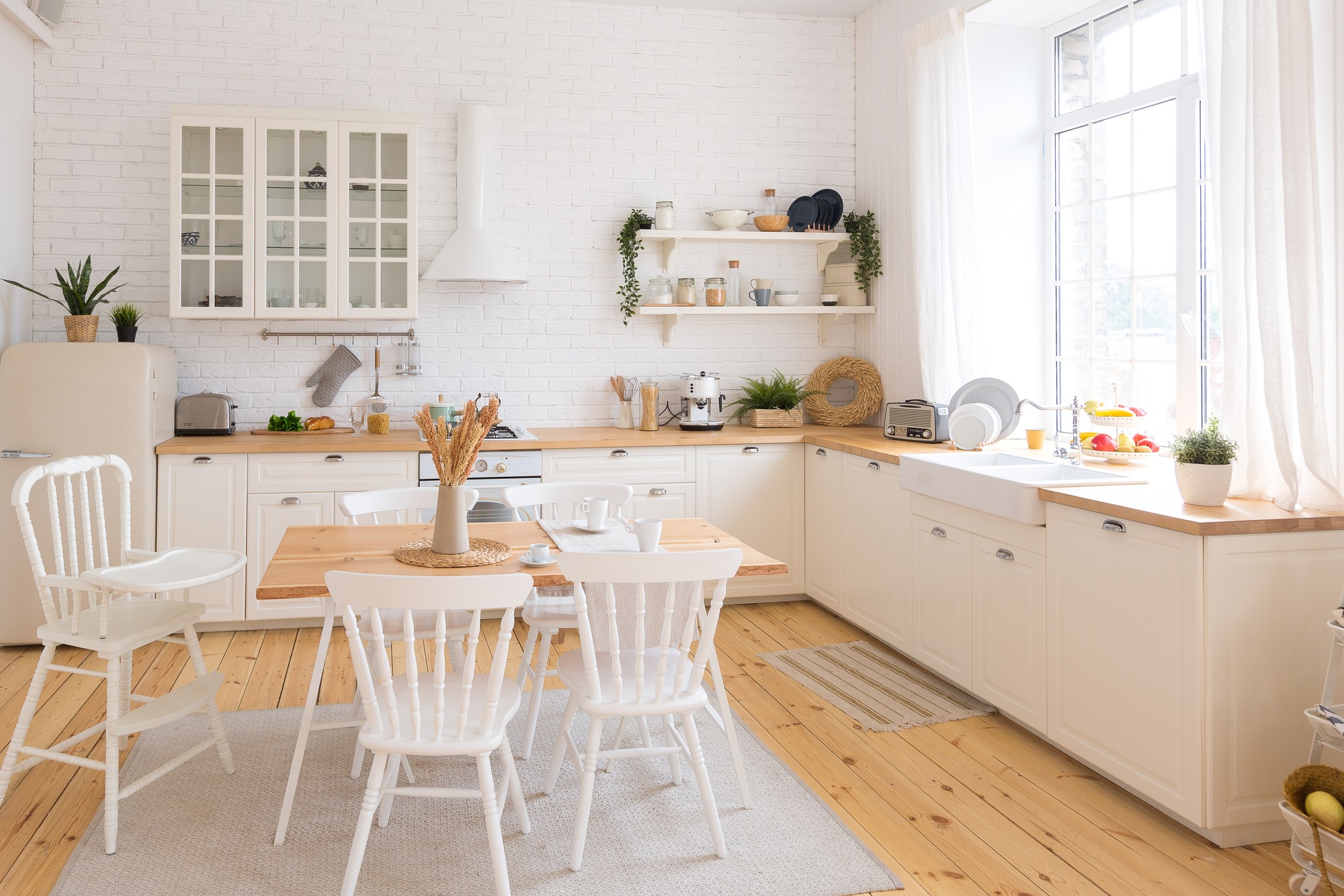 Jasna kuchnia w stylu angielskim z drewnianym blatem, białymi szafkami i ceglaną ścianą, uzupełniona drewnianym stołem i białymi krzesłami, oświetlona naturalnym światłem.