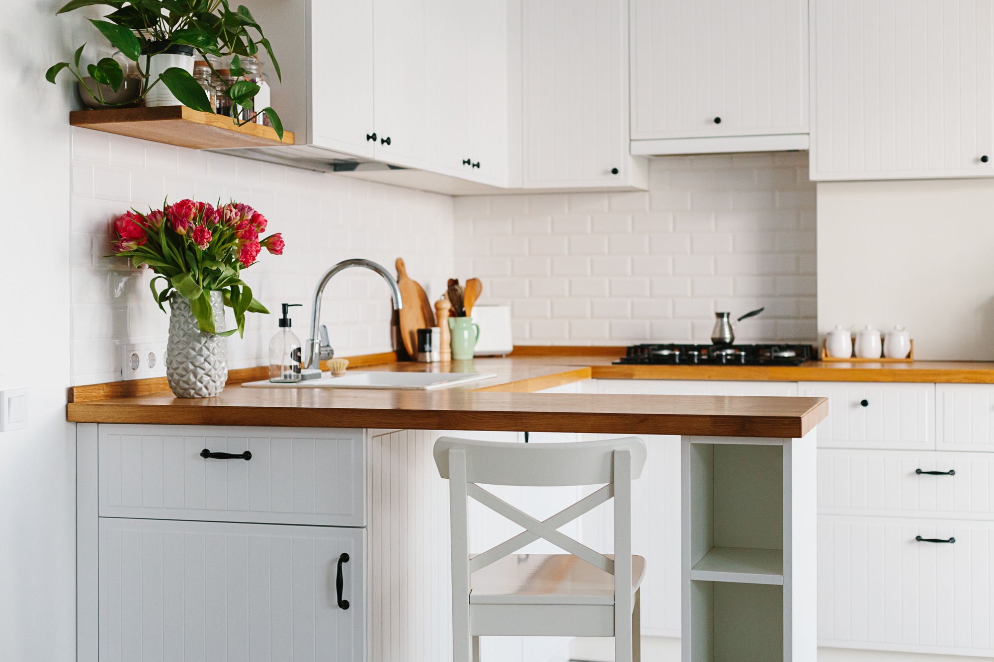 Jasna kuchnia w stylu skandynawskim z drewnianym blatem, białymi szafkami z frezowanymi frontami i czarnymi uchwytami, ozdobiona bukietem czerwonych tulipanów w wazonie o fakturze ananasa.
