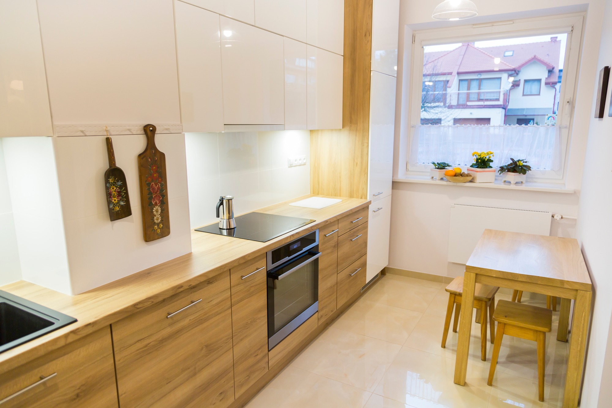 Jasna kuchnia z drewnianymi blatami i białymi szafkami, ozdobiona ludowymi deskami do krojenia, z widokiem na dom przez okno.