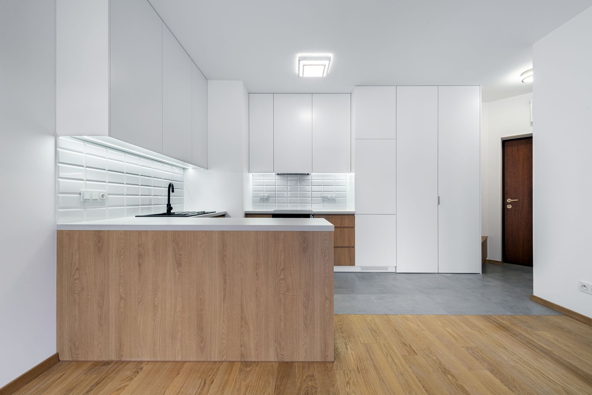 Nowoczesna, minimalistyczna kuchnia z białymi szafkami i drewnianym blatem wyspowym, podświetlona ledami, z widocznym fragmentem parkietu i szarej podłogi w tle