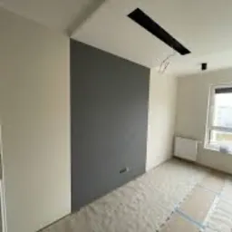 Pomalowane na szaro fragmenty ściany w jasnym pomieszczeniu, podłoga zabezpieczona folią malarską, widoczne gniazdka elektryczne i okno.