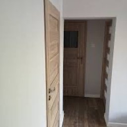 BUDMAR - Jasne, drewniane drzwi wewnętrzne z ozdobnymi panelami, zamontowane w świeżo pomalowanym na szaro pomieszczeniu z podłogą imitującą drewno.