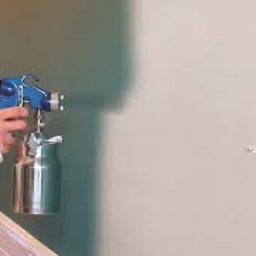 BUDMAR - Pracownik w białym kombinezonie maluje ścianę natryskowo, używając pistoletu lakierniczego z metalowym zbiornikiem, widoczne delikatne rozpryski farby na ścianie.