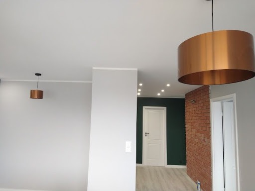 Wnętrze mieszkania po remoncie z szarą ścianą, ceglaną ścianką działową, białymi drzwiami i wiszącymi miedzianymi lampami.