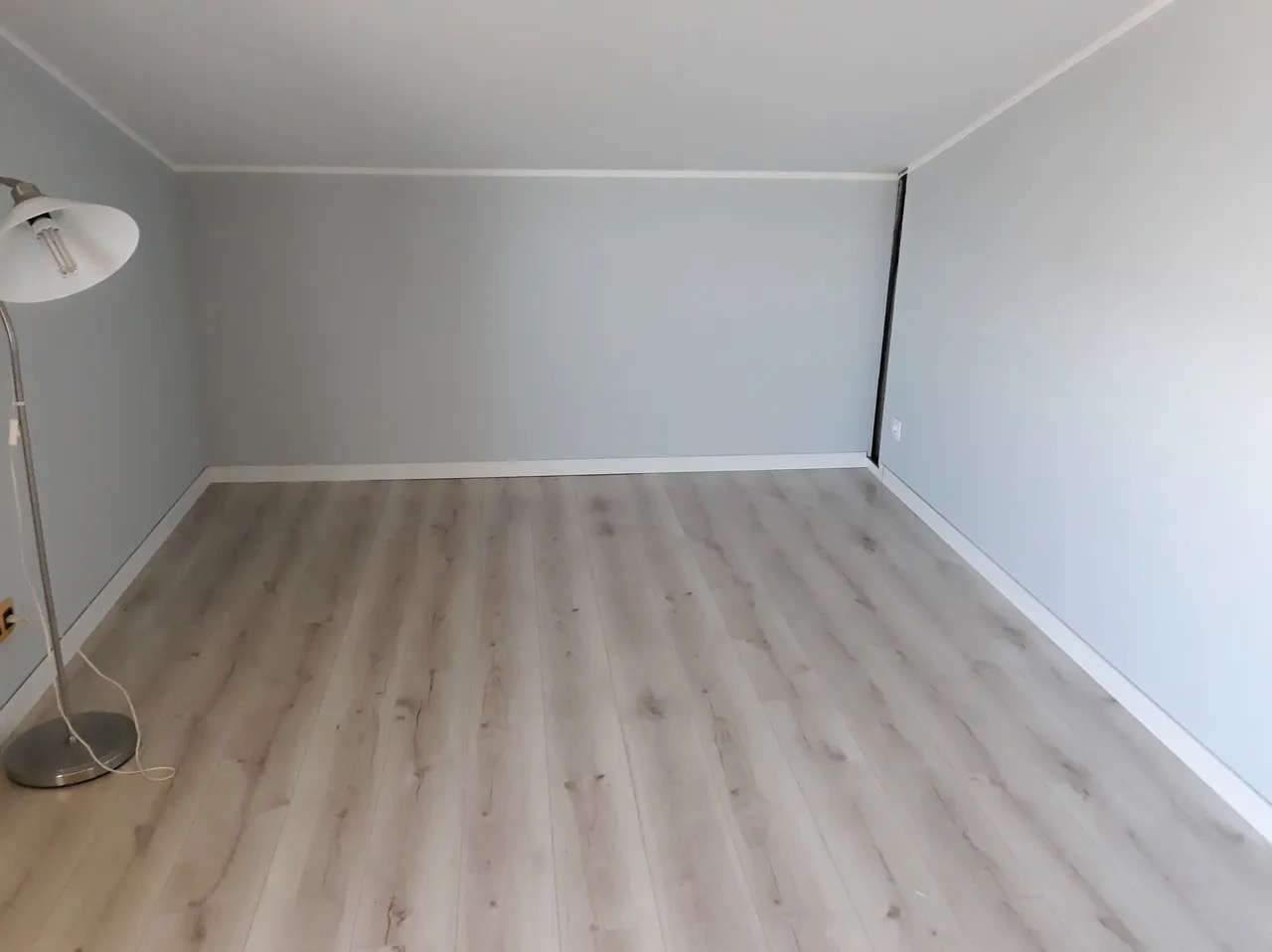 Pusty, świeżo odnowiony pokój z panelami podłogowymi w odcieniach jasnego drewna i szarymi ścianami, oświetlony lampą podłogową z metalowym kloszem.