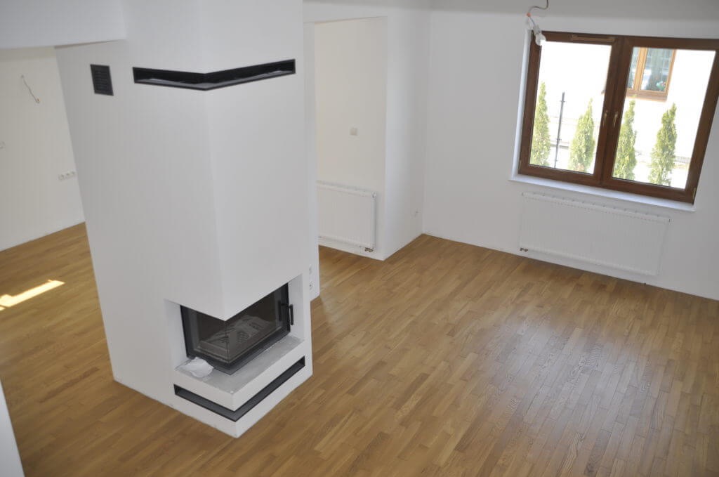 Jasne, puste wnętrze z drewnianą podłogą parkietową, białymi ścianami, grzejnikami i kominkiem z czarnymi akcentami, widok na zewnątrz przez okno z czterema drzewami.