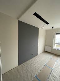 Pomalowane na szaro fragmenty ściany w jasnym pomieszczeniu, podłoga zabezpieczona folią malarską, widoczne gniazdka elektryczne i okno.