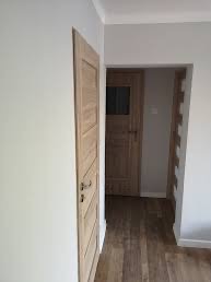 Jasne, drewniane drzwi wewnętrzne z ozdobnymi panelami, zamontowane w świeżo pomalowanym na szaro pomieszczeniu z podłogą imitującą drewno.