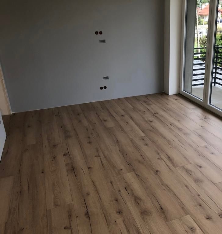 Jasne panele podłogowe z widoczną strukturą drewna w nowym pokoju z szarą ścianą i przygotowanymi gniazdkami elektrycznymi, balkon z czarną barierką w tle.