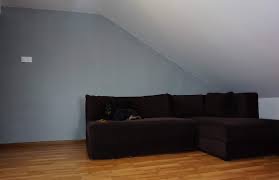 Szary pokój na poddaszu z brązowym narożnikiem i drewnianą podłogą. Widoczny ukośny sufit i minimalistyczny design.