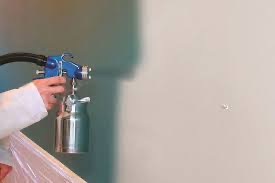 Pracownik w białym kombinezonie maluje ścianę natryskowo, używając pistoletu lakierniczego z metalowym zbiornikiem, widoczne delikatne rozpryski farby na ścianie.