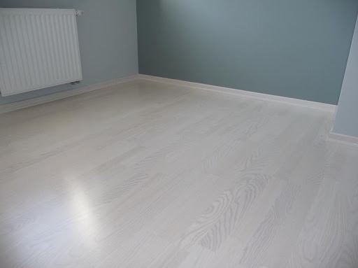 Jasne, bielone panele podłogowe ułożone w pustym pokoju z szarobłękitnymi ścianami i białym grzejnikiem.