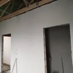 Surowe ściany w trakcie prac wykończeniowych, widoczne dwa otwory drzwiowe, ściany pokryte szarą warstwą, drewniana konstrukcja dachu z widocznym zwisającym kablem.