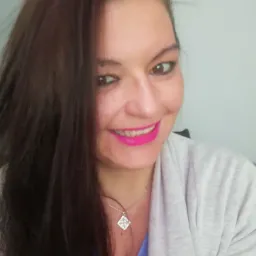 Uśmiechnięta kobieta z długimi ciemnymi włosami, różową szminką i srebrnym naszyjnikiem z zawieszką w kształcie rombu, ubrana w szary sweter, pozuje na jasnym tle.