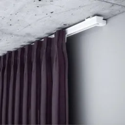 Zasłony w kolorze ciemnej śliwki zawieszone na białym, sufitowym karniszu w minimalistycznym wnętrzu.  Widoczna faktura surowego betonu na suficie.