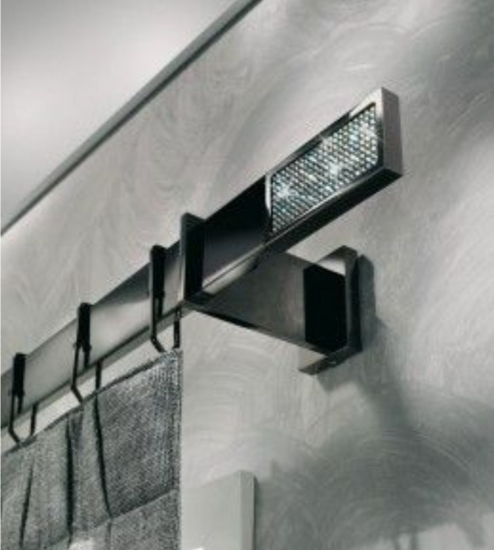 Metalowy karnisz z dekoracyjnym elementem z kryształkami, zawieszony na ścianie o fakturze betonu, z fragmentem szarej zasłony na haczykach.