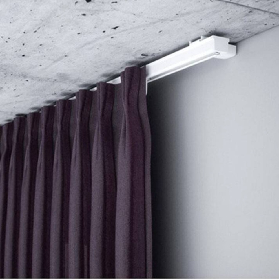 Zasłony w kolorze ciemnej śliwki zawieszone na białym, sufitowym karniszu w minimalistycznym wnętrzu.  Widoczna faktura surowego betonu na suficie.