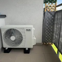 Zewnętrzna jednostka klimatyzacji AUX DC Inverter na balkonie z szarymi płytkami, widoczna puszka jedzenia dla kota i osłona balkonowa