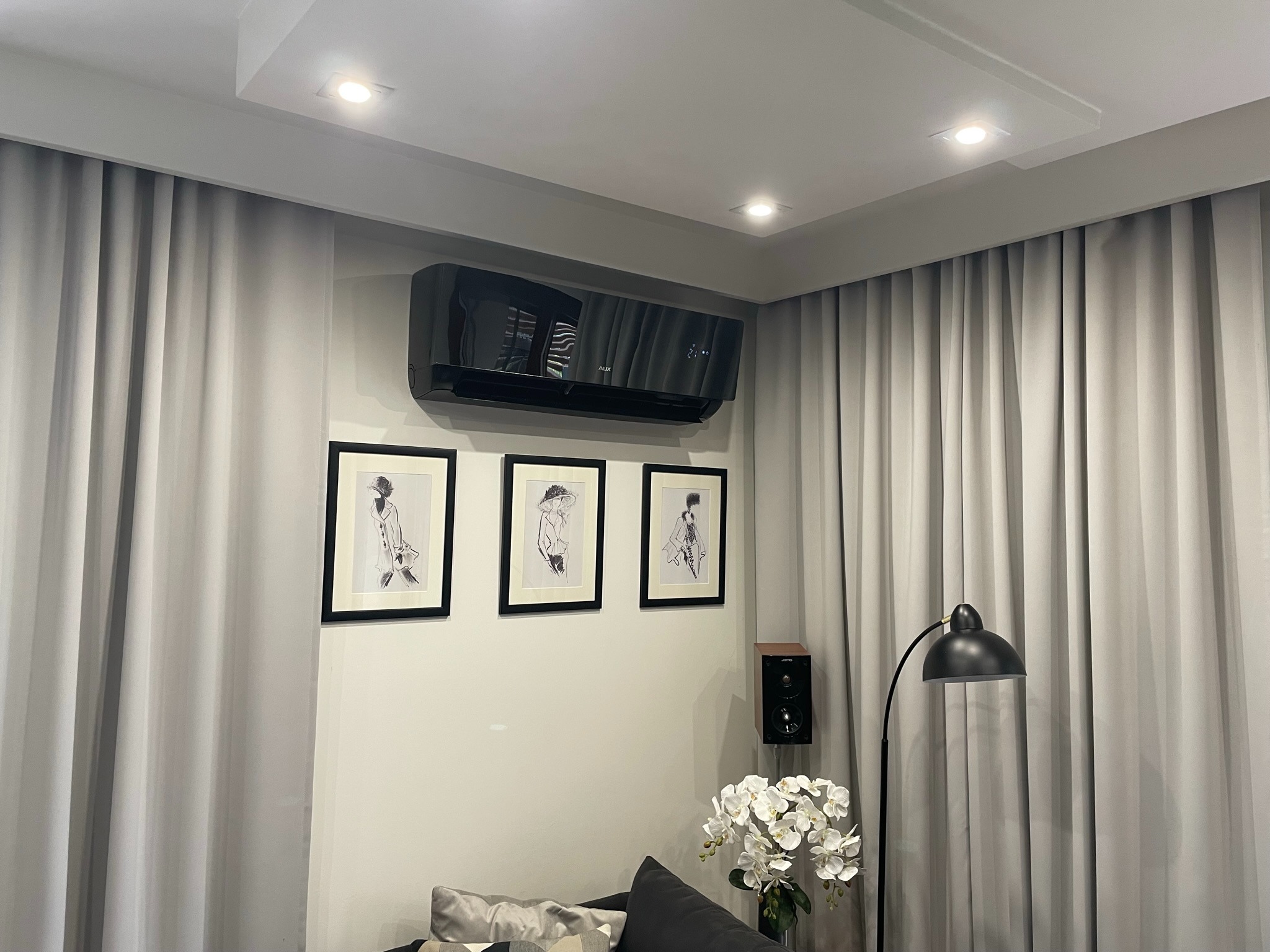 Czarny klimatyzator ścienny marki AUX w nowoczesnym salonie z szarymi zasłonami, oświetleniem punktowym i trzema czarno-białymi obrazami w ramkach.
