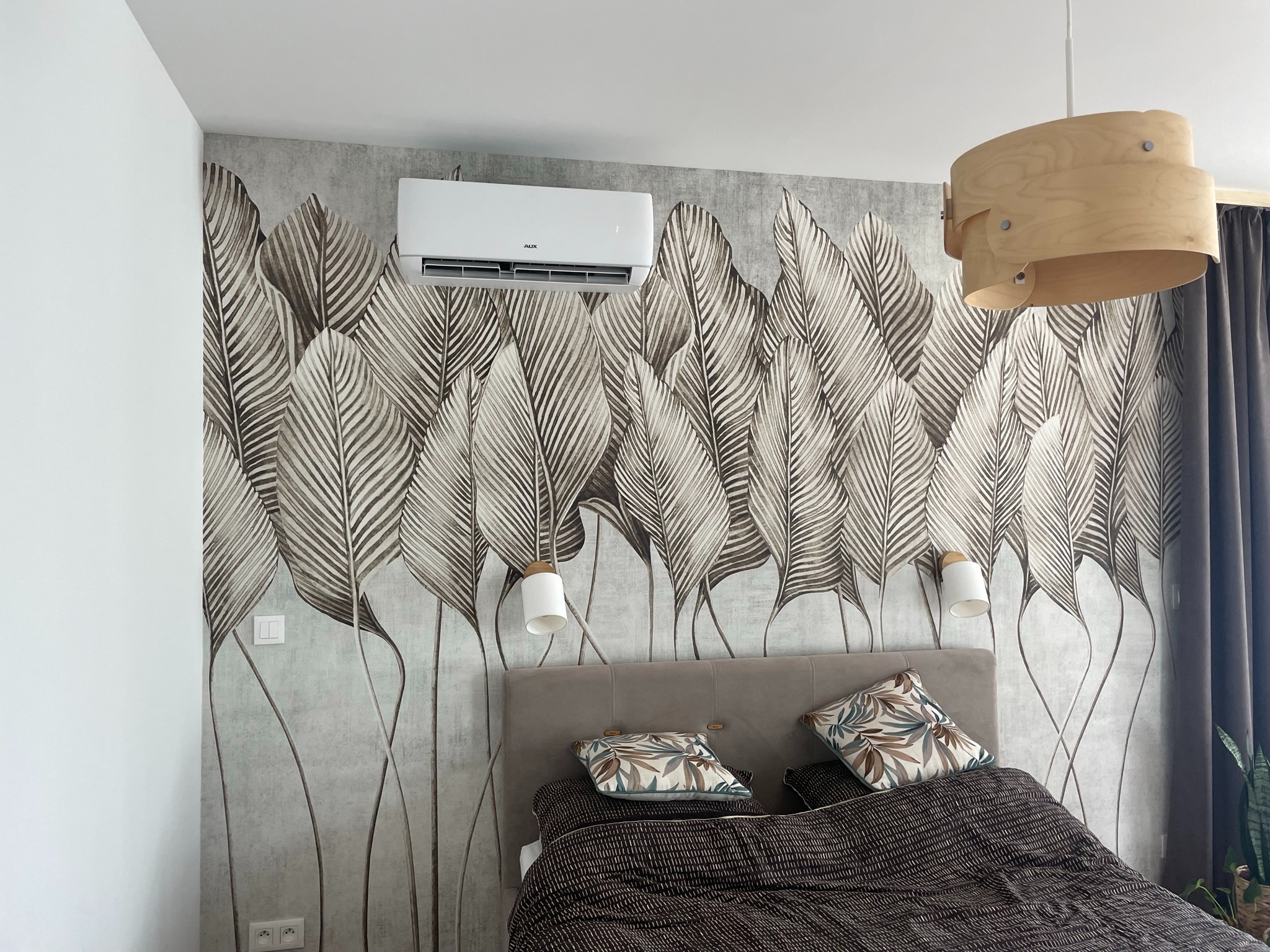 Biała klimatyzacja AUX zamontowana na ścianie z tapetą w liście nad łóżkiem w sypialni.