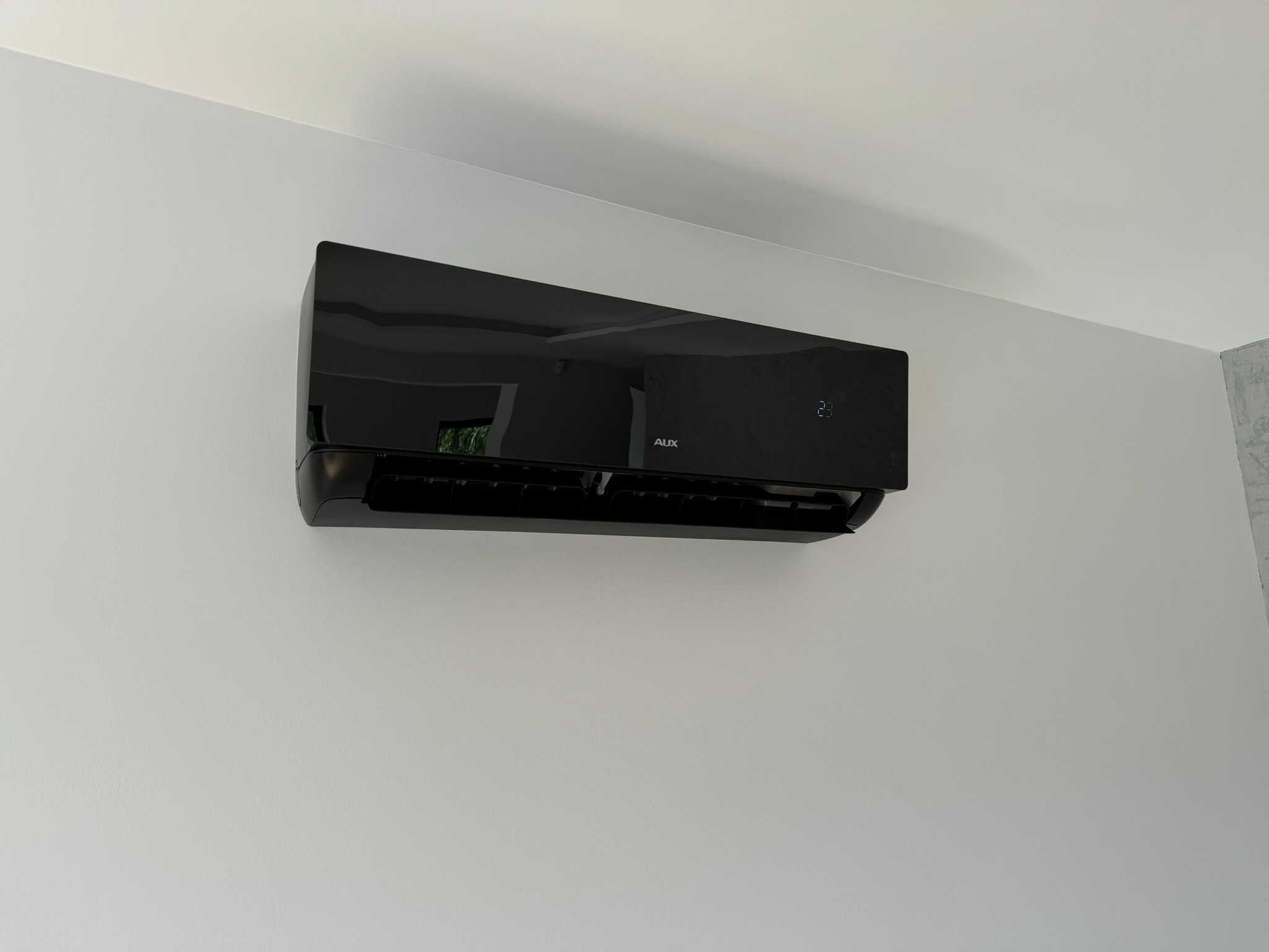 Czarny klimatyzator AUX zamontowany na białej ścianie, widoczny wyświetlacz z temperaturą 23 stopnie.