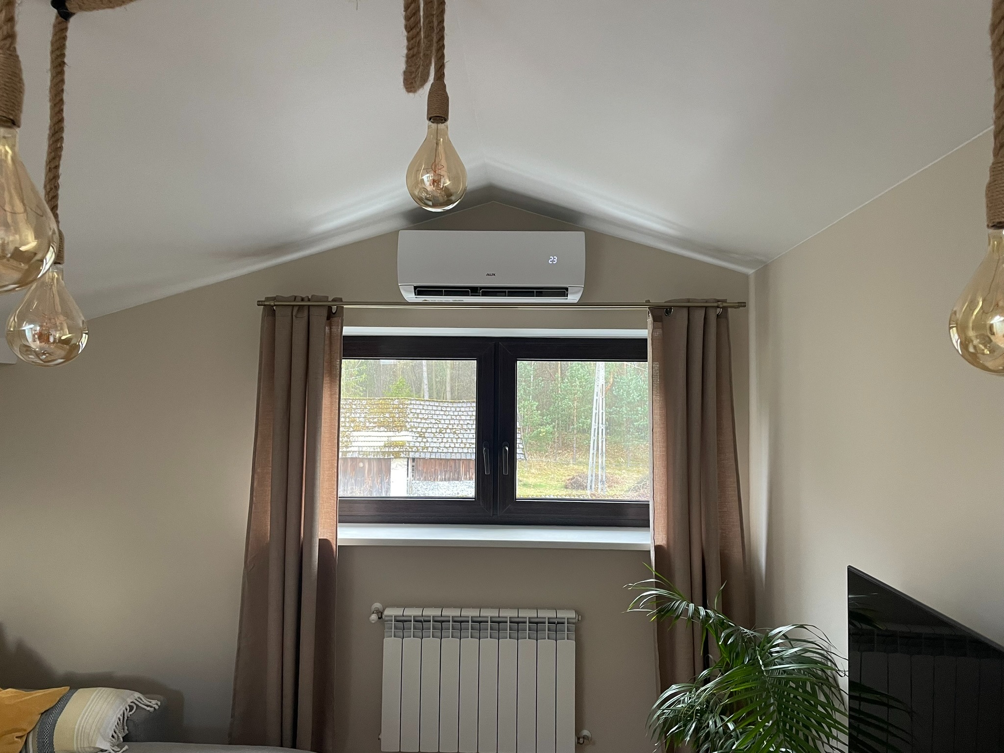 Biały klimatyzator ścienny z wyświetlaczem 23 stopni, zamontowany nad oknem z brązowymi ramami i zasłonami w pokoju z beżowymi ścianami, wiszącymi lampami na sznurach i grzejnikiem pod oknem.