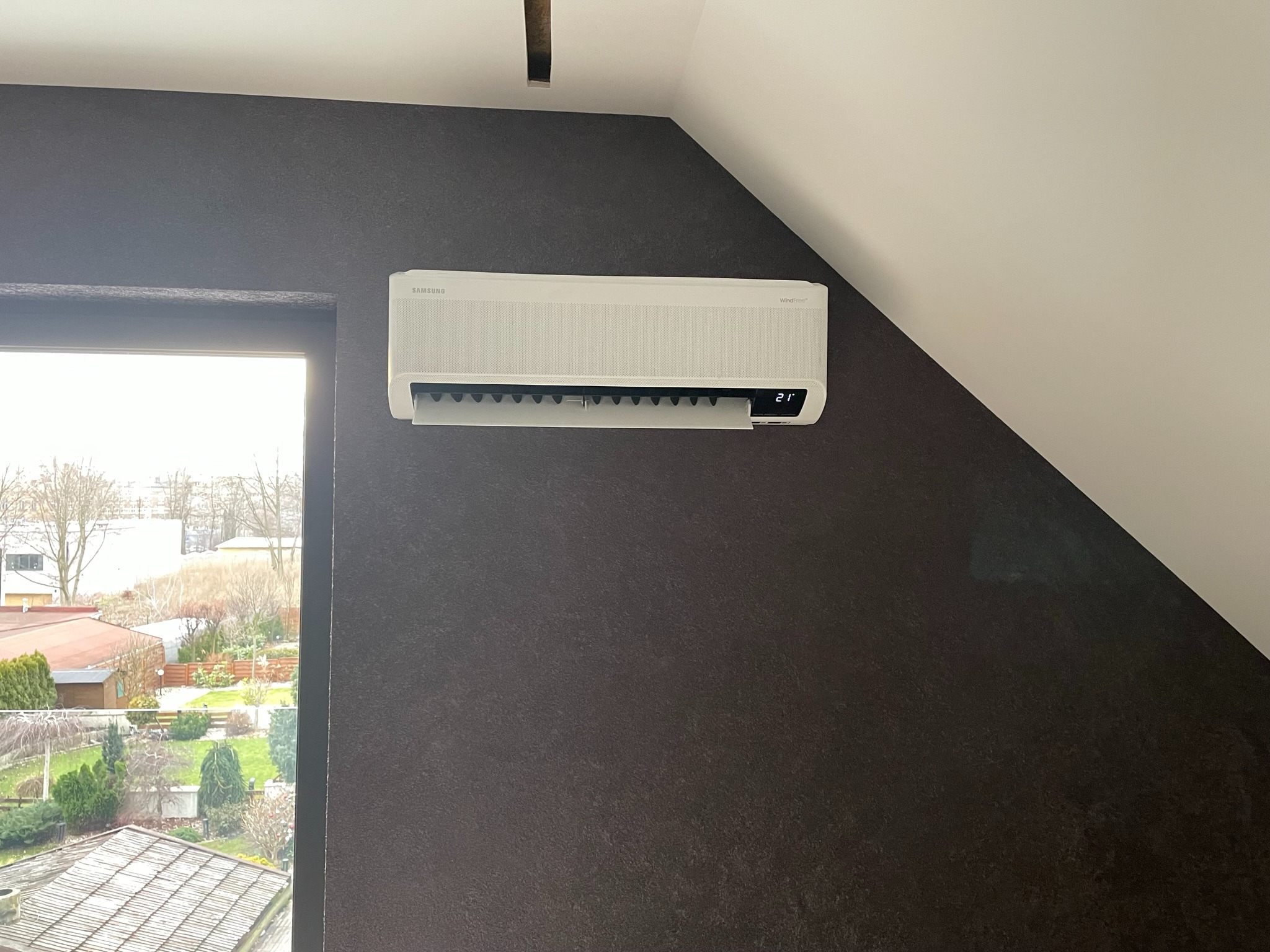 Biały klimatyzator Samsung WindFree zamontowany na ciemnoszarej ścianie w pokoju na poddaszu, widok z okna na ogród.