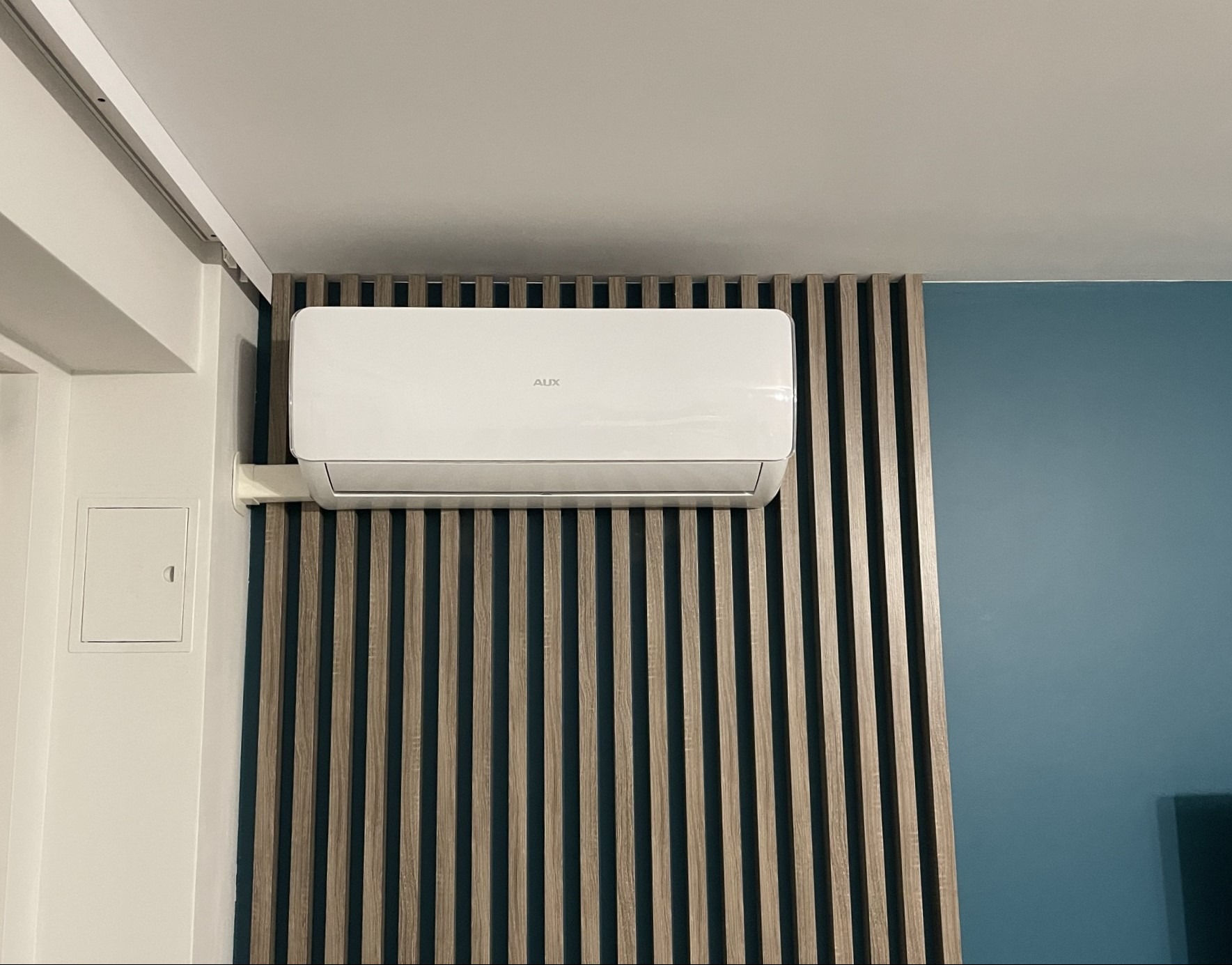 Biała jednostka klimatyzacyjna marki AUX zamontowana na ścianie z dekoracyjnymi, pionowymi panelami drewnopodobnymi, obok białej ściany z małą skrzynką elektryczną i niebieskiej ściany.