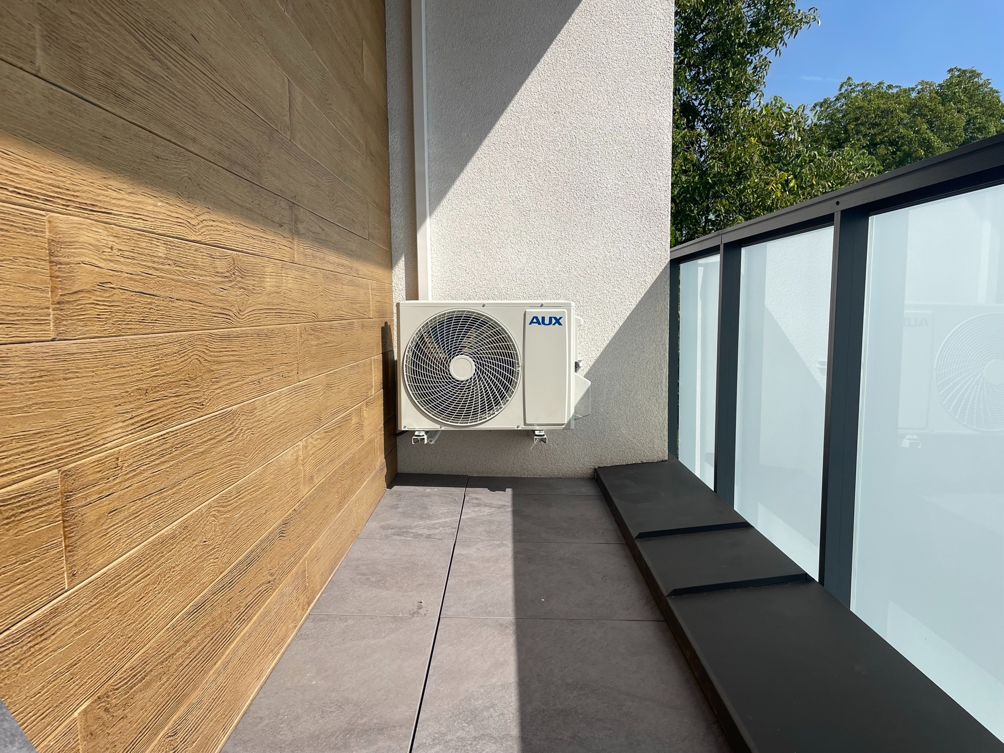 Biała jednostka zewnętrzna klimatyzatora marki AUX zamontowana na elewacji z drewnopodobnych paneli i białego tynku, widok z zewnątrz balkonu z szklaną balustradą.