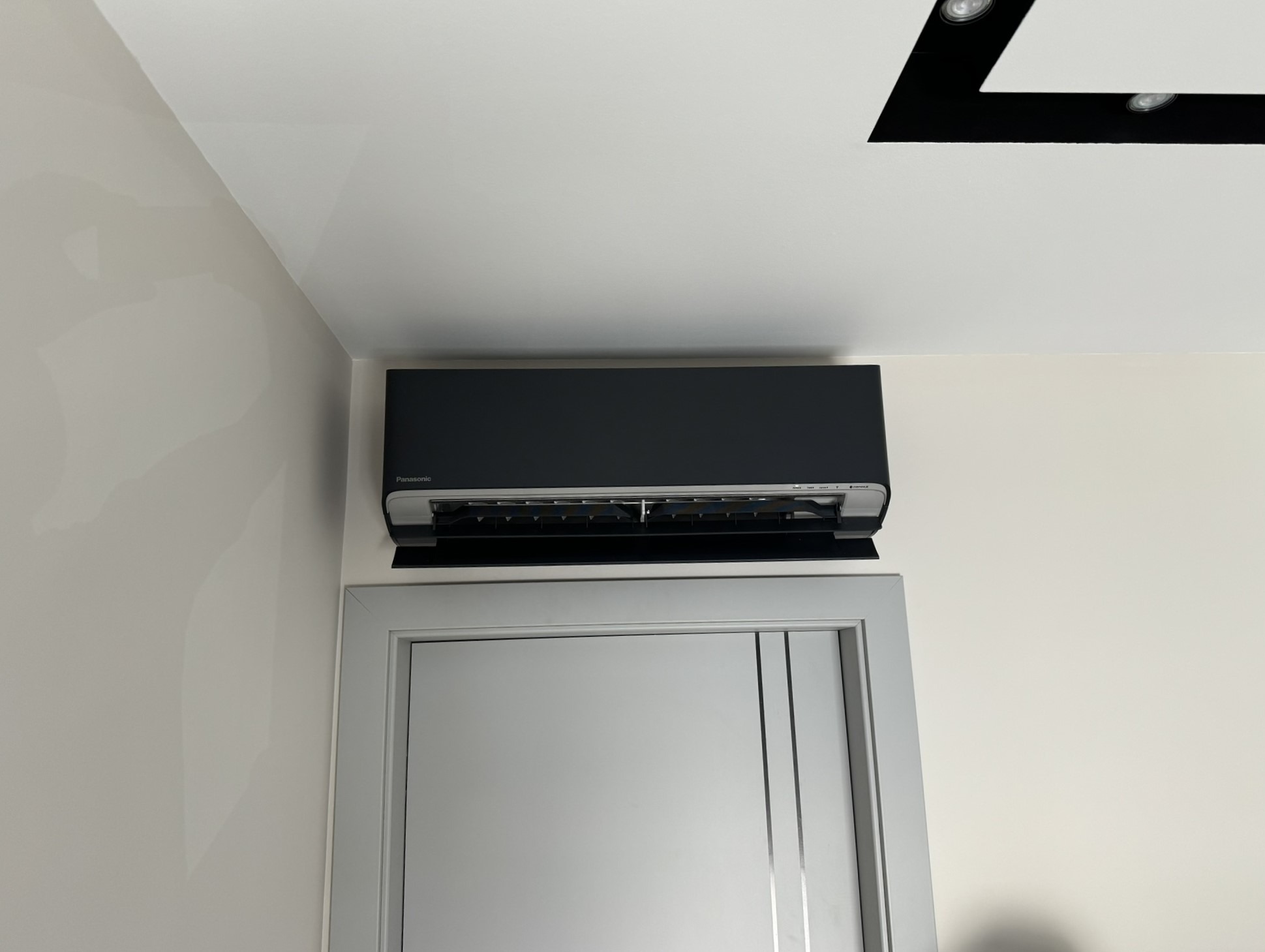 Czarny klimatyzator Panasonic zamontowany nad szarymi drzwiami w pomieszczeniu z kremowymi ścianami i białym sufitem z czarną wstawką i oświetleniem punktowym.