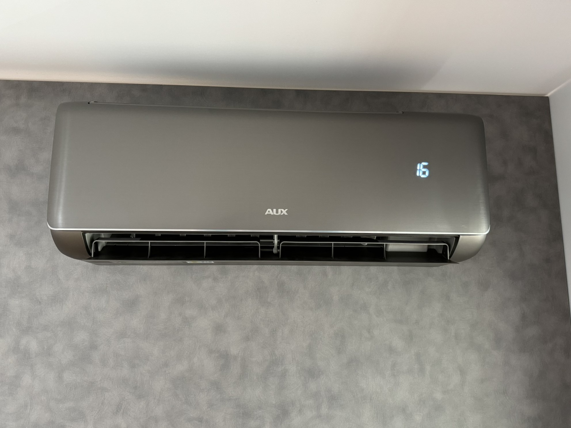 Szary klimatyzator AUX na ścianie z dekoracyjnym tynkiem, wyświetlacz pokazuje temperaturę 16 stopni.