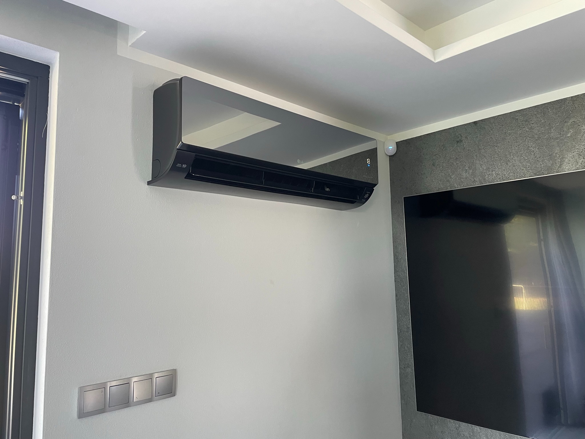 Czarny klimatyzator ścienny LG z wyświetlaczem temperatury, zamontowany w nowoczesnym wnętrzu obok szarej ściany z telewizorem i zestawu włączników.