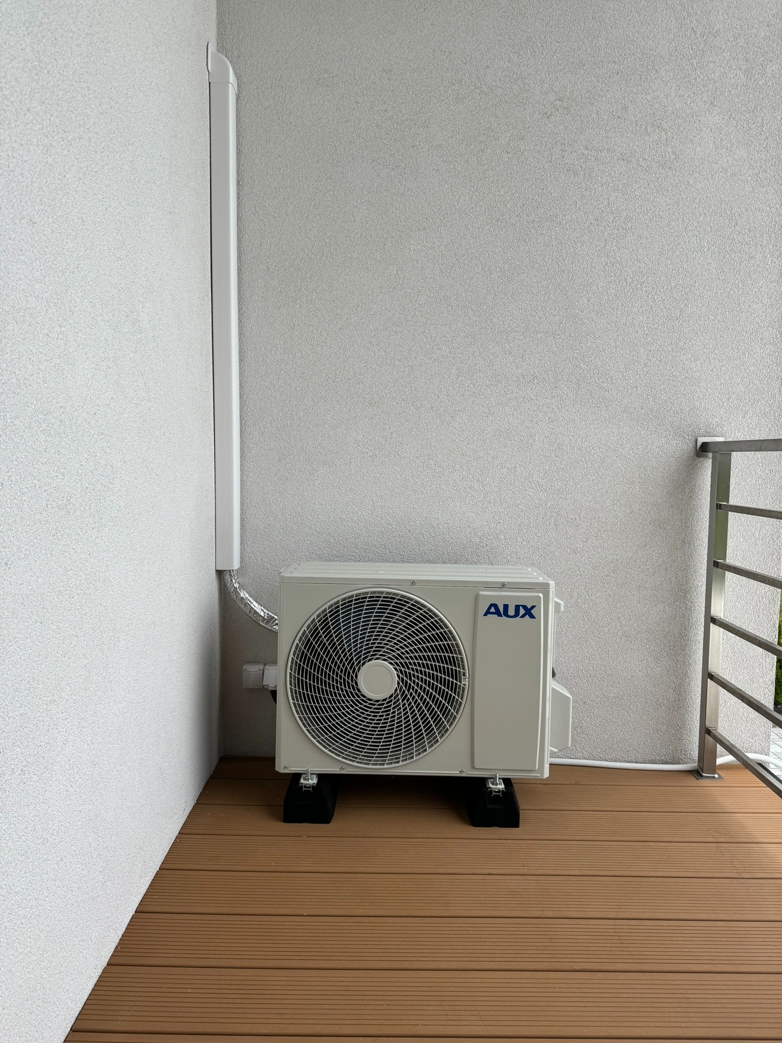 Zewnętrzna jednostka klimatyzacji marki AUX zamontowana na balkonie z brązową podłogą kompozytową, z widocznymi przewodami i osłoną instalacji przy ścianie.